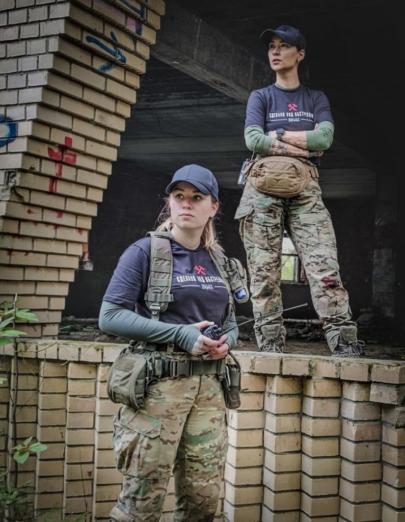Две девушки в тактической экипировке, камуфляжных брюках Multicam и черных футболках на фоне кирпичных руин